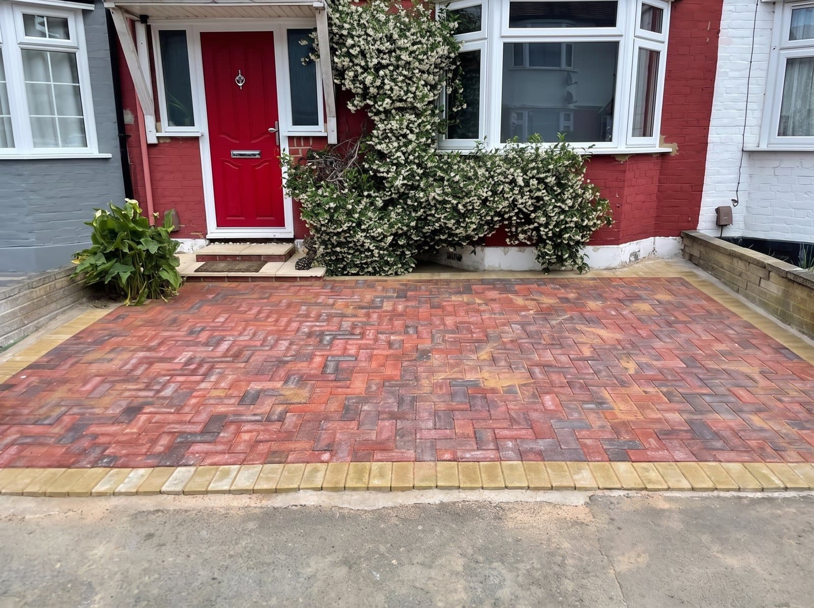 A front garden paved with red and grey blocks in front of a house with a red door